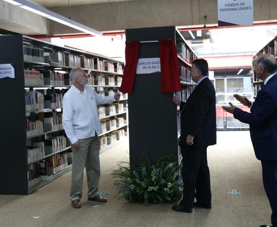 3 Personas mirando una placa metalica, que se encuentra montada en un estante que contiene los libros donados.